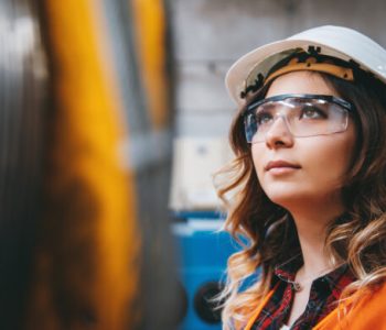 Portrait of young beautiful engineer woman working in factory building. Portrait of young businesswoman with white helmet looking up and seen from the industrial steel cable reel for crane in factory warehouse. She is testing and working with winding wire and cable drum accessories metal wire spool reel wear resistance.