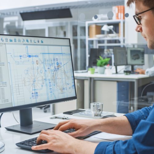 Over the Shoulder Shot of Engineer Working with CAD Software on Desktop Computer, Screen Shows Technical Drafts and Drawings. In the Background Engineering Facility Specialising on Industrial Design Over the Shoulder Shot of Engineer Working with CAD Software on Desktop Computer, Screen Shows Technical Drafts and Drawings. In the Background Engineering Facility Specialising on Industrial Design