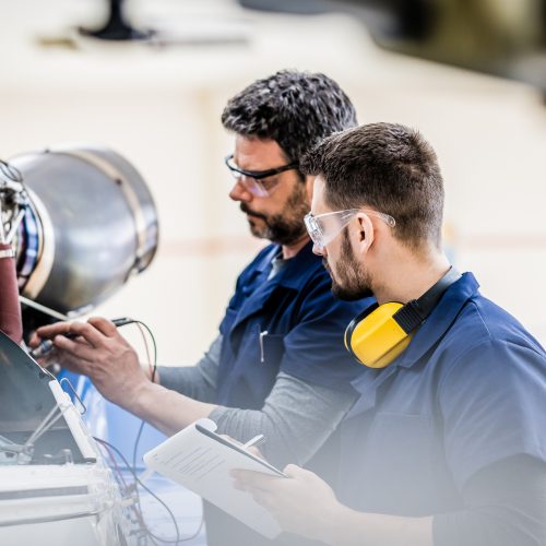 Aviation mechanic teaching coworker examining engine, side view Aviation mechanic teaching coworker examining engine, side view. Aircraft hangar workers in protective equipment holding clipboard, medium shot